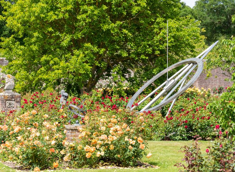 Large ornamental sundial sculpture set among vibrant rose beds and lush greenery in a formal garden setting.